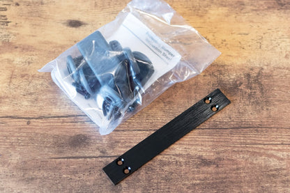 A black metal mounting bracket with pre-drilled holes and a clear plastic bag containing black hardware, displayed on a wooden tabletop.