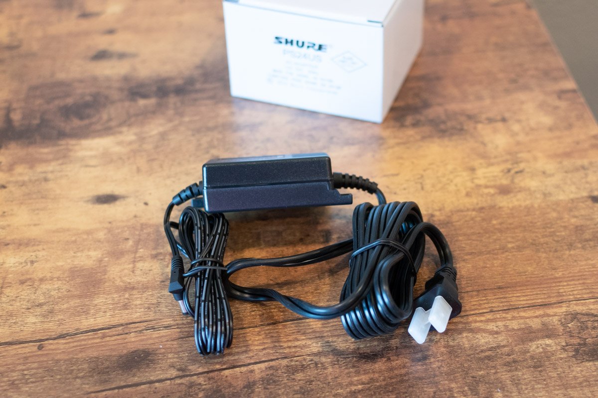 Black power adapter with a coiled cable on a wooden surface, with a white box labeled 'SHURE' in the background.
